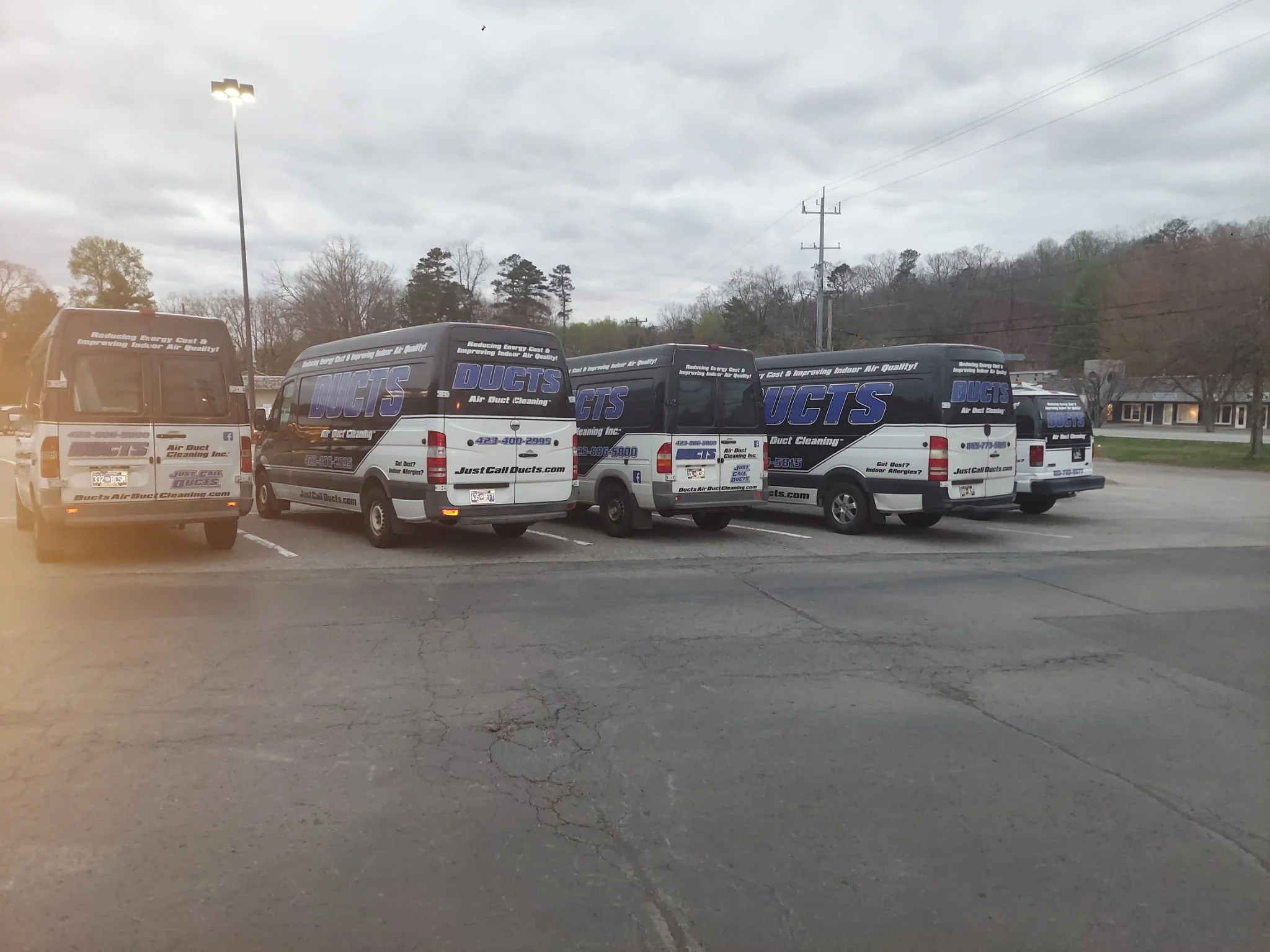 Five company vans parked in a parking lot, all branded with 'Just Call DUCTS' for air duct cleaning services. The vans feature the company's website, contact number, and logos.