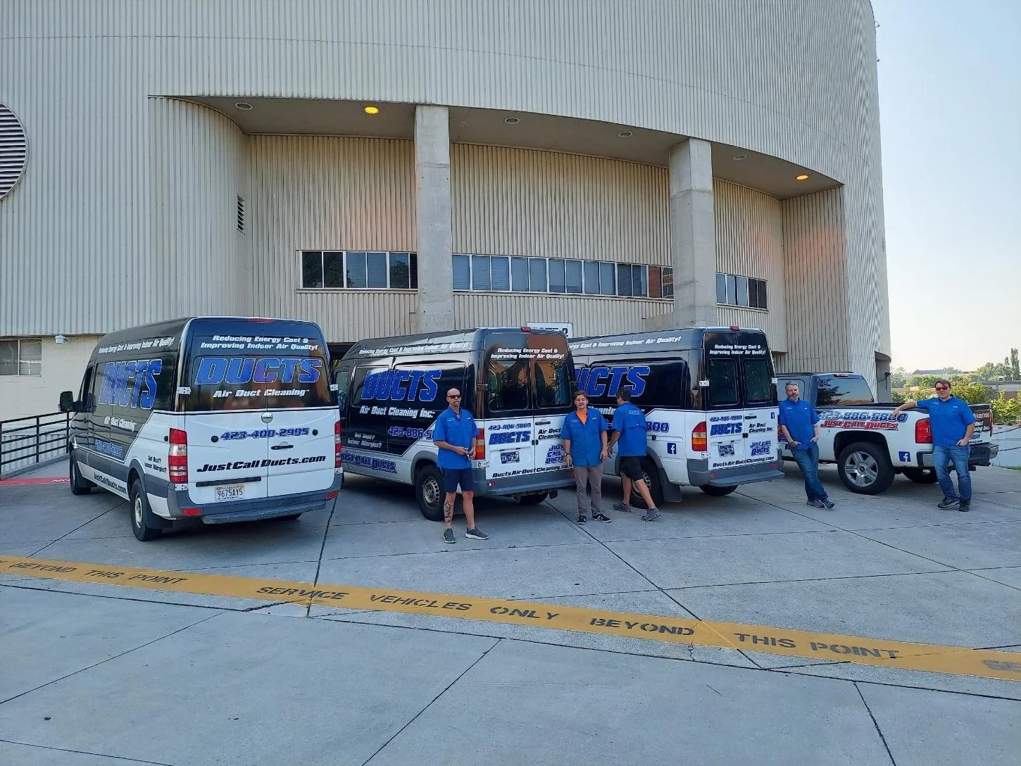 Five men in blue shirts standing and leaning against trucks with DUCTS branding, parked in front of a modern building, with yellow service vehicles tape on the ground.
