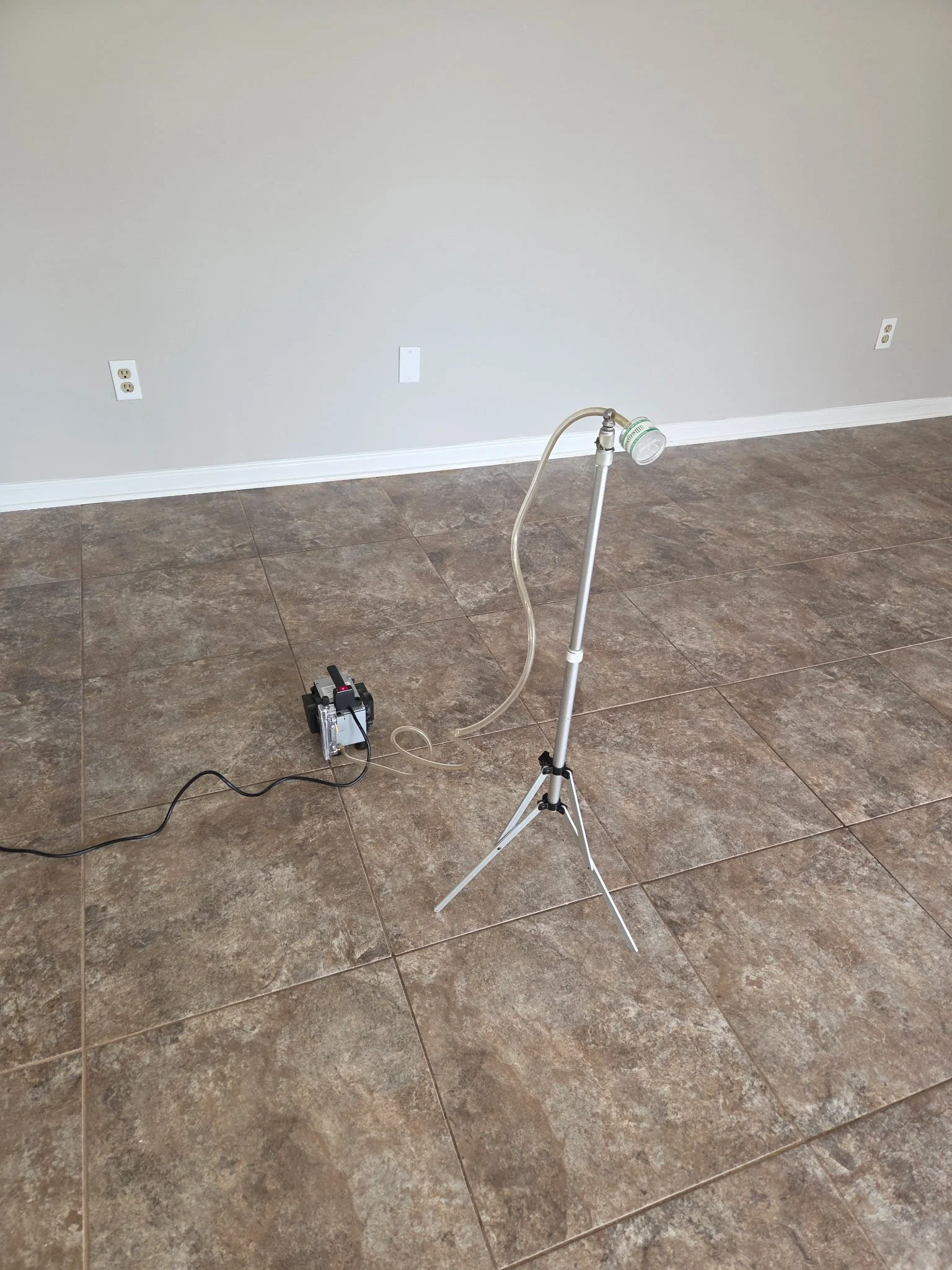 A scientific setup with a tripod, a vacuum pump, and a tube leading to a container, on a tiled floor against a plain wall with electrical outlets.