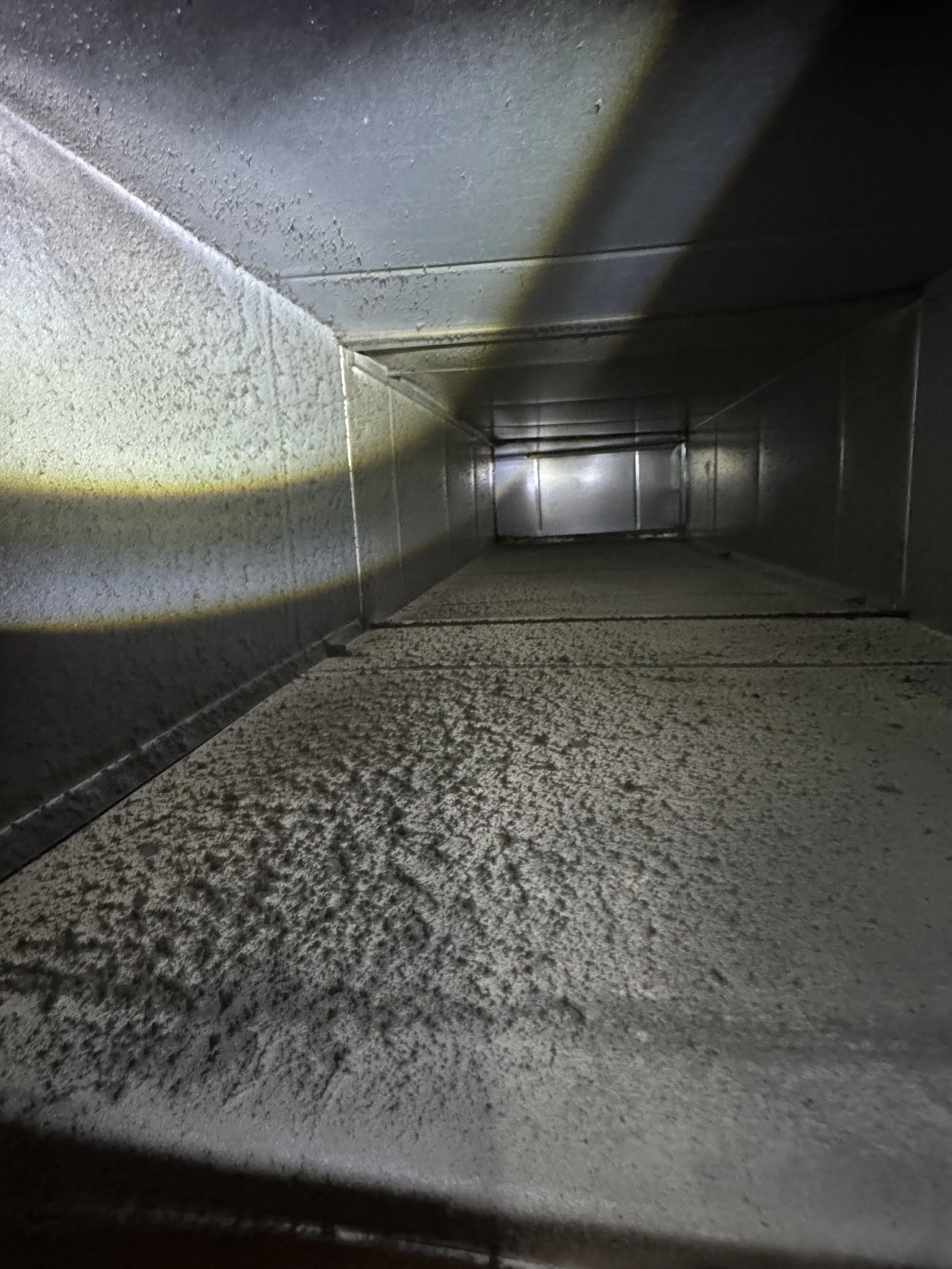 Looking into a narrow metallic duct or vent with a textured surface, illuminated by a bright flashlight or light source.