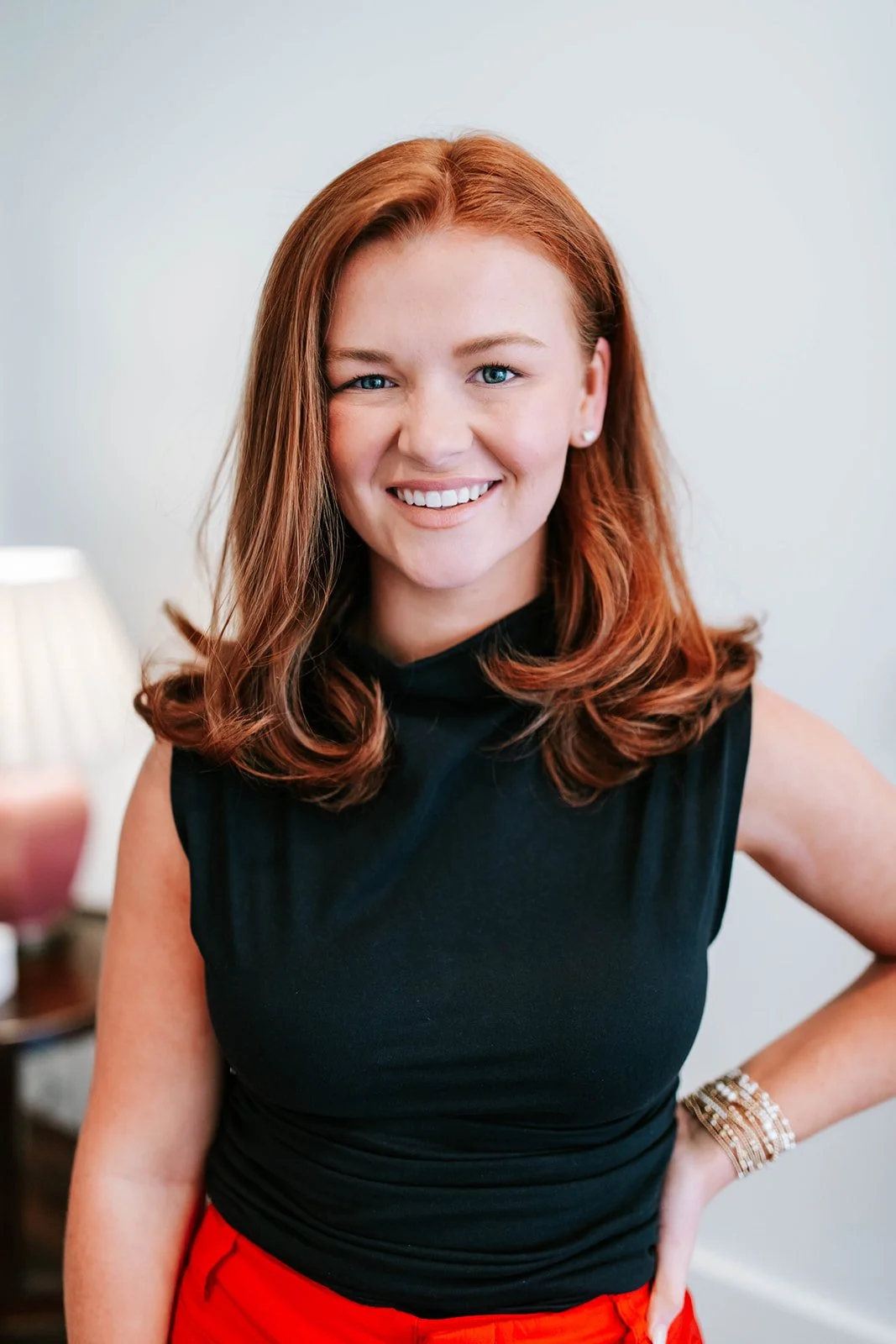 A young woman with red hair smiling in a black sleeveless top and red pants, standing indoors.
