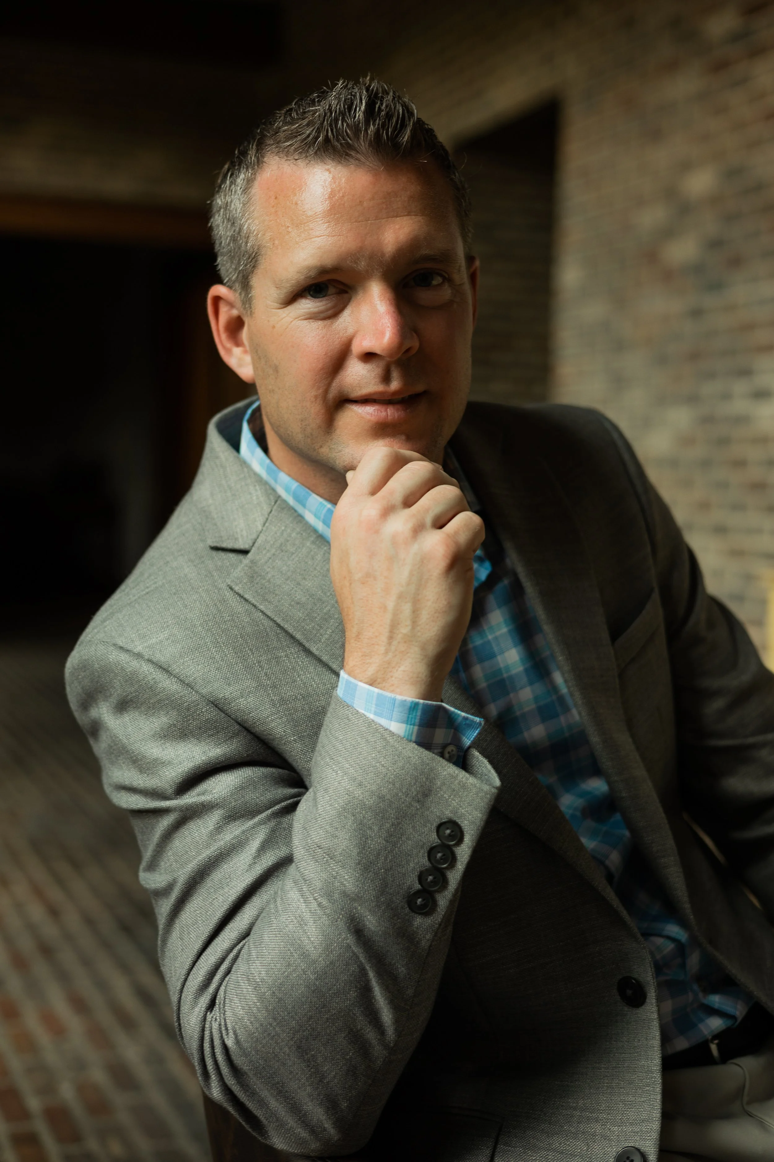 A man in a grey suit with a blue checkered shirt, sitting in a room with a brick wall background, resting his chin on his hand and looking at the camera.