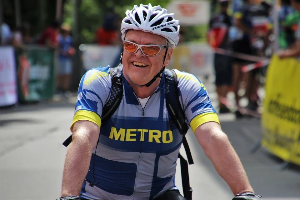 A smiling cyclist wearing a white helmet and sunglasses, dressed in a blue and yellow METRO cycling jersey, riding outdoors during a race or event with onlookers and barricades in the background.