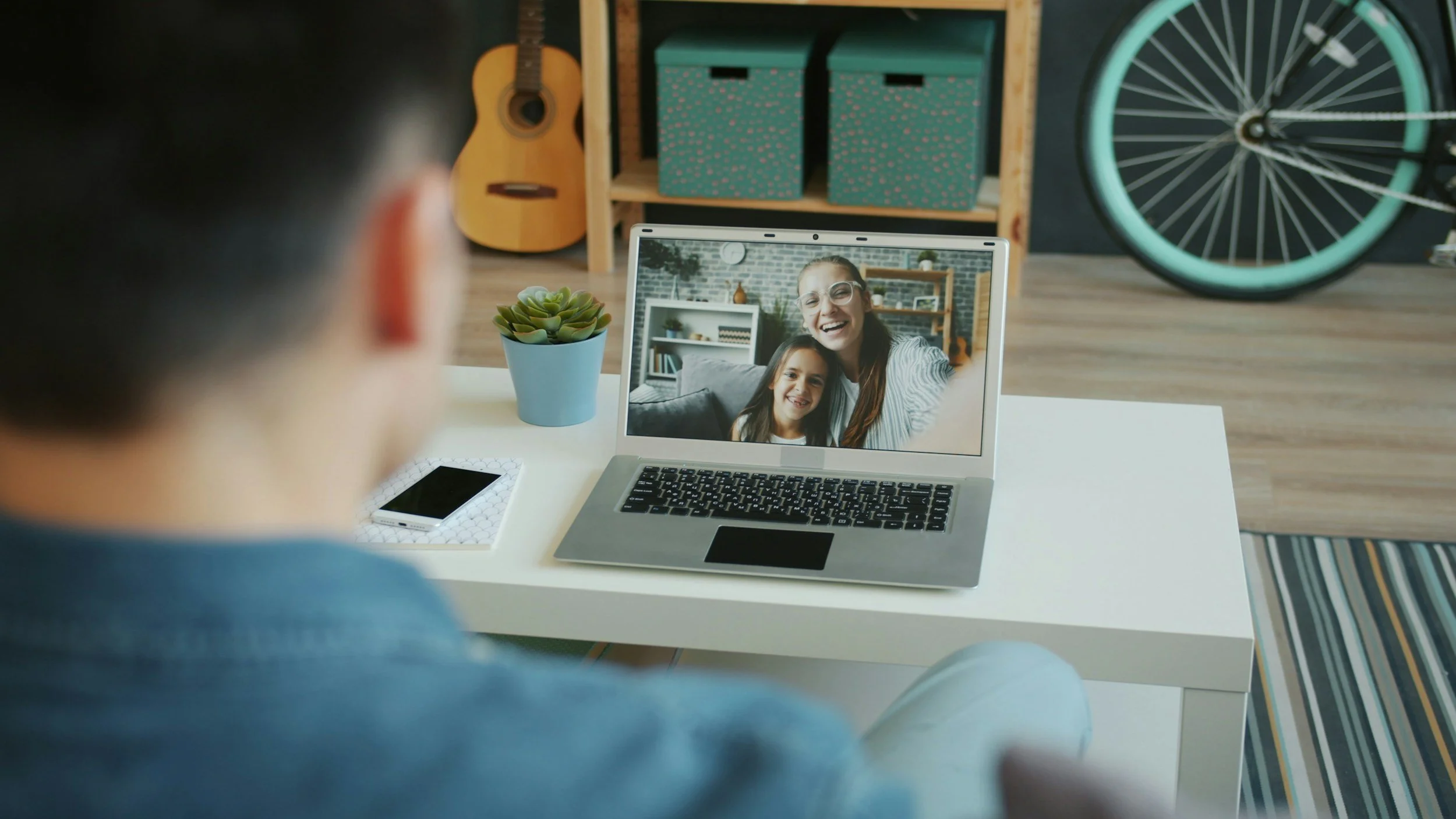 Person on a video call with a woman and a girl displayed on a laptop screen, sitting at a white table with a smartphone, a plant, and a notebook nearby in a cozy living room.
