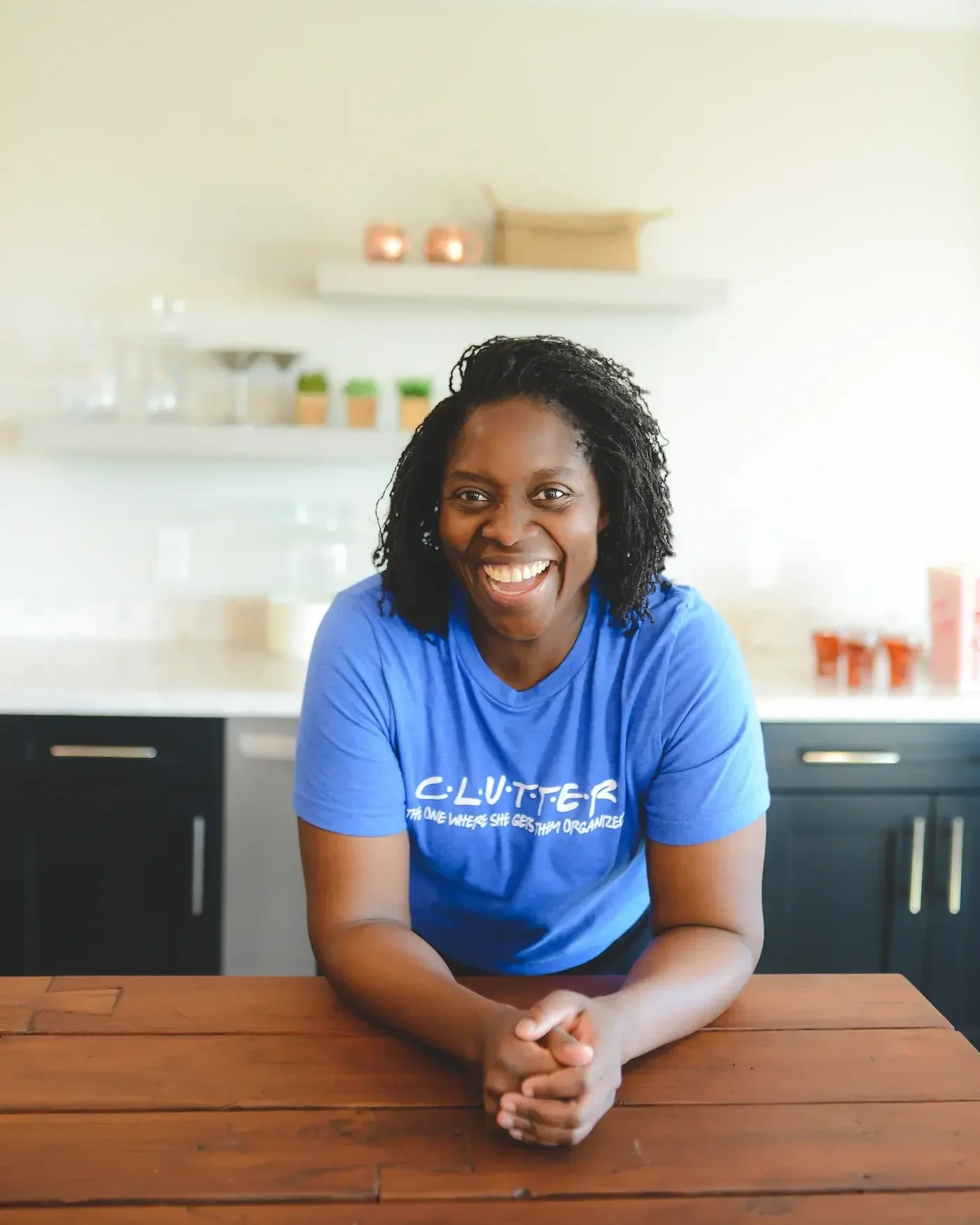 A smiling woman with curly black hair, wearing a blue T-shirt, leaning on a wooden table in a modern kitchen.