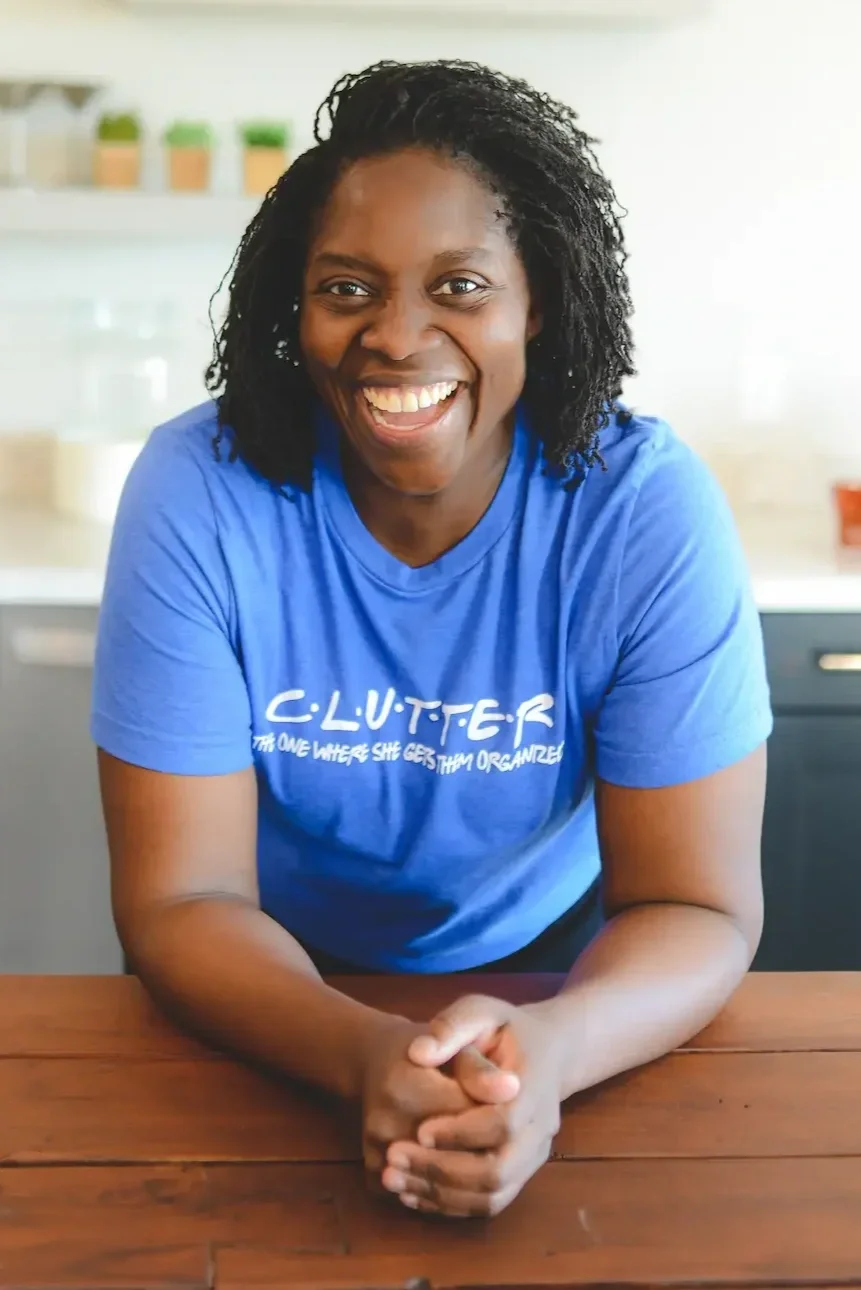 A woman with dark curly hair wearing a blue t-shirt that says 'CLUTTER THE ONE WHERE SHE GETS THEM ORGANIZED' smiling and leaning on a wooden surface in a bright room with a kitchen background.