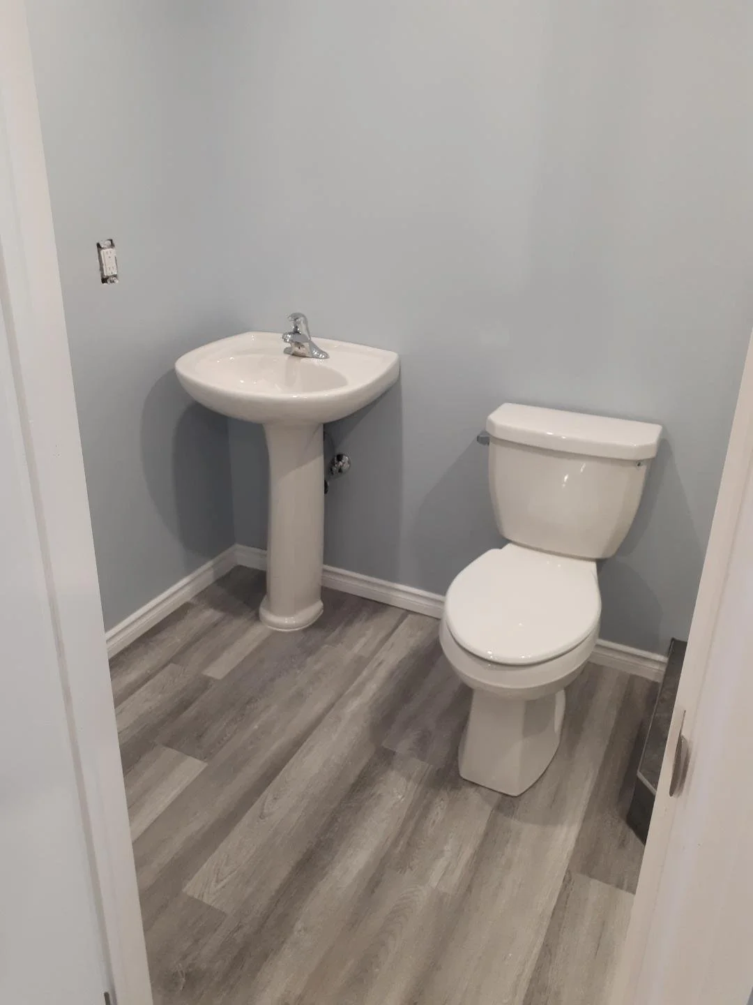 Small bathroom with light gray walls, a white toilet, and a small white pedestal sink. The bathroom has gray wood-look flooring and is unfurnished.