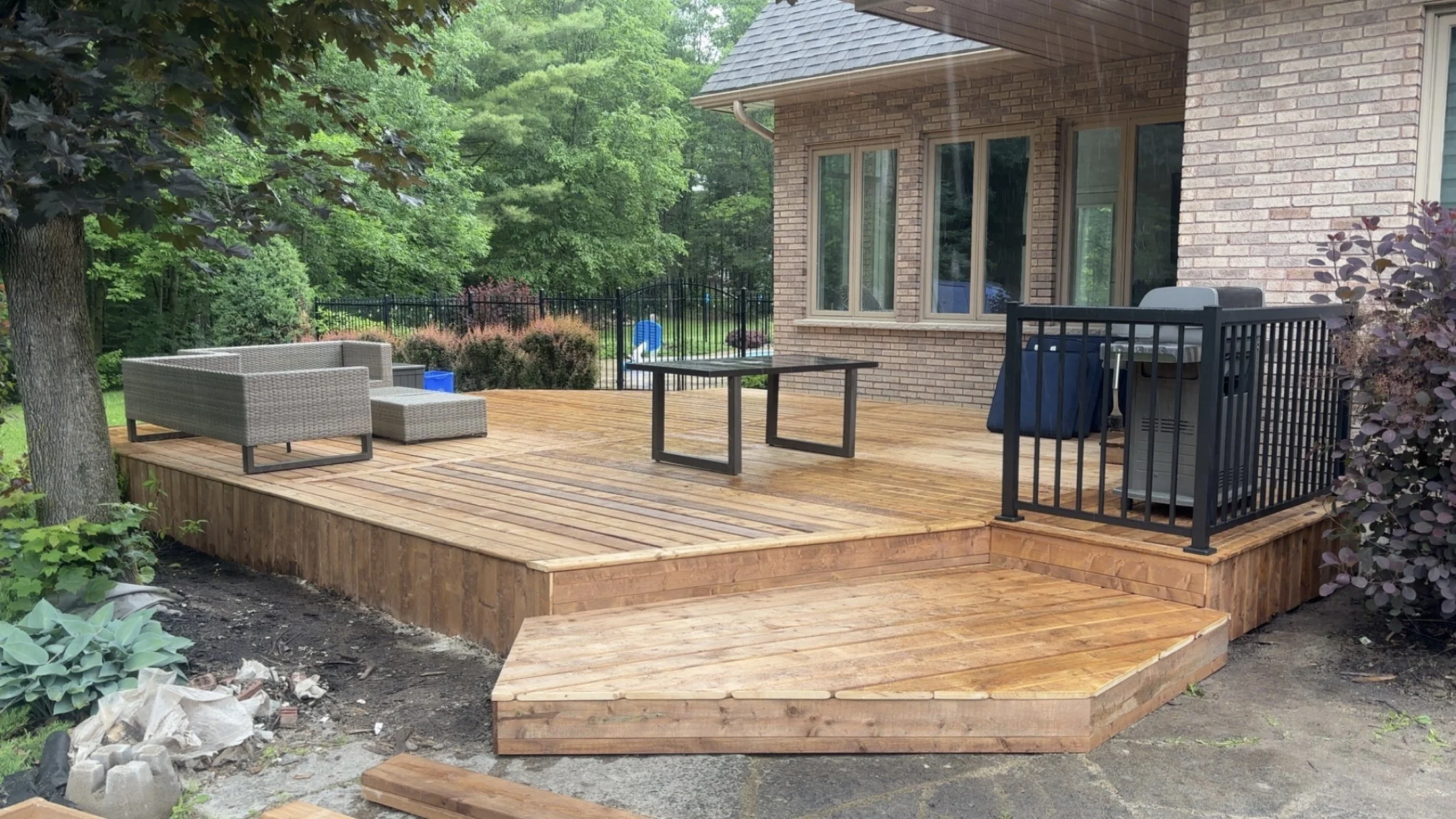 New wooden deck attached to a brick house, with outdoor furniture including a wicker sofa, a black metal table, and a barbecue grill, surrounded by green trees and bushes.