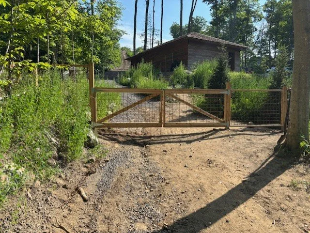 Wooden gate on dirt path leading to a rustic house in a wooded area with trees and bushes.