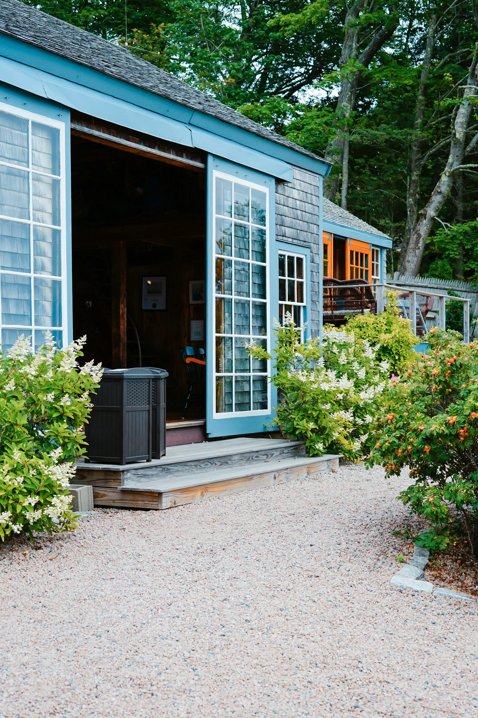 A small blue wooden cottage with open French doors, surrounded by greenery and flowering bushes, with a gravel pathway leading to the entrance.