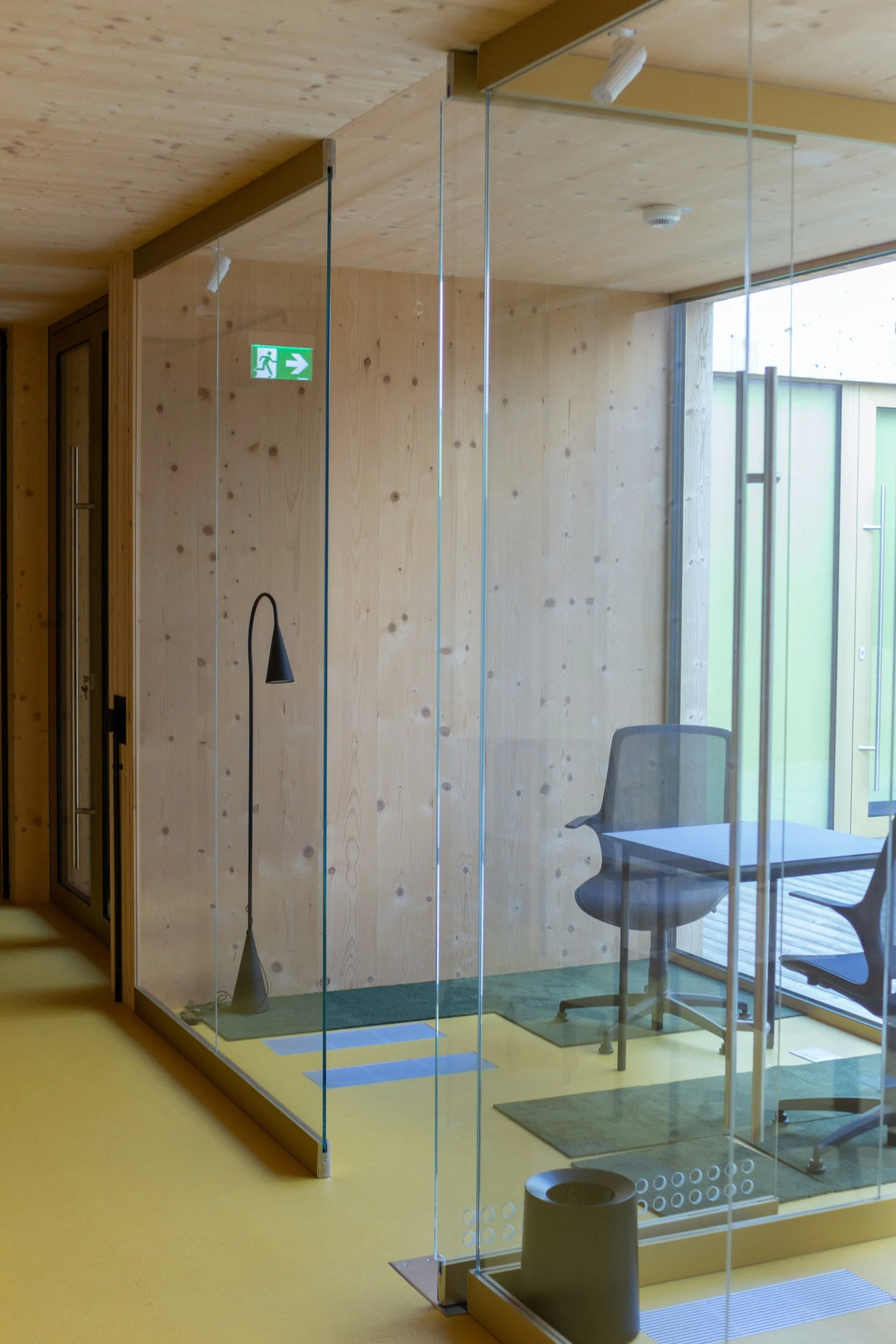 An interior office space with a glass partition, wooden walls, a yellow floor, a black curved lamp, and a chair, with a large window.