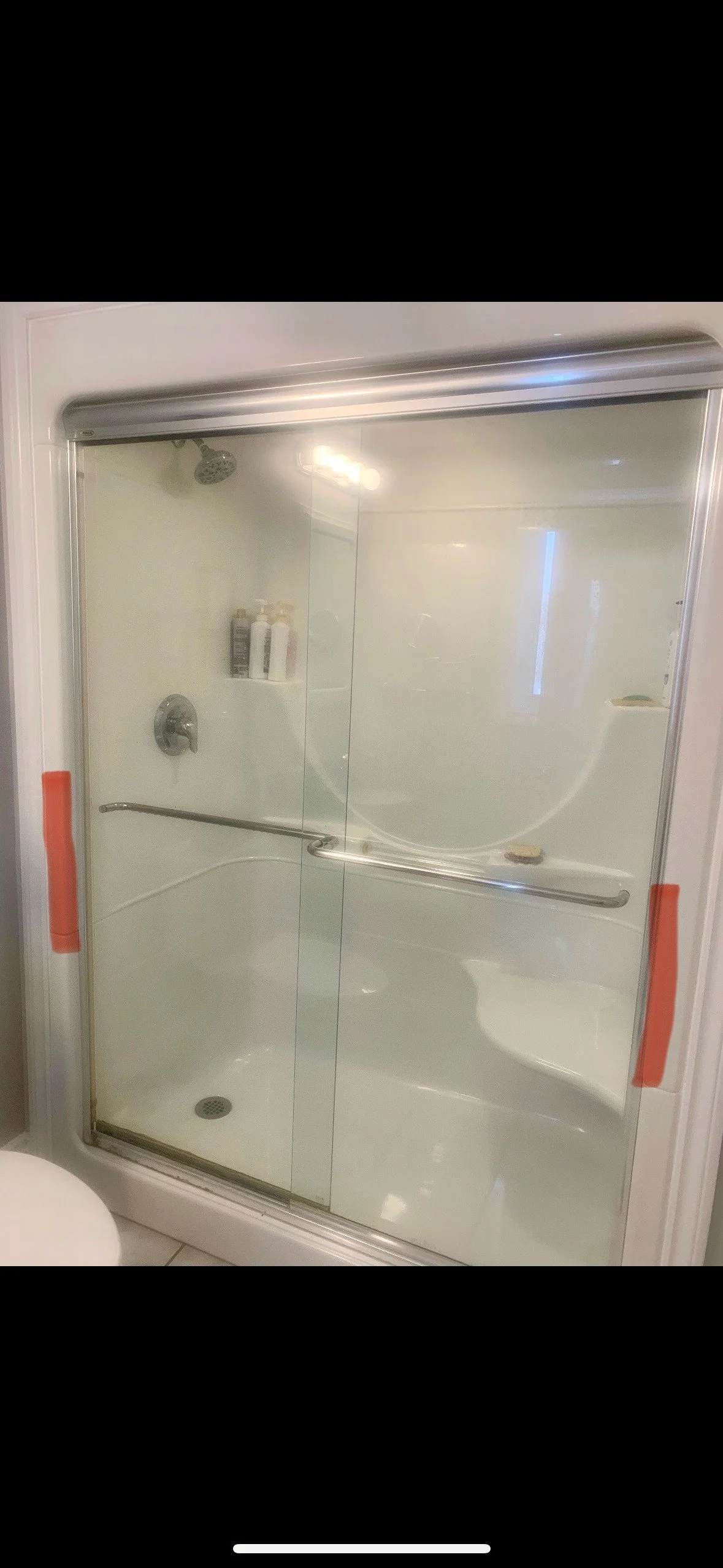 Enclosed shower with sliding glass doors, a handrail, a wall-mounted showerhead, and a small shelf with bottles inside.