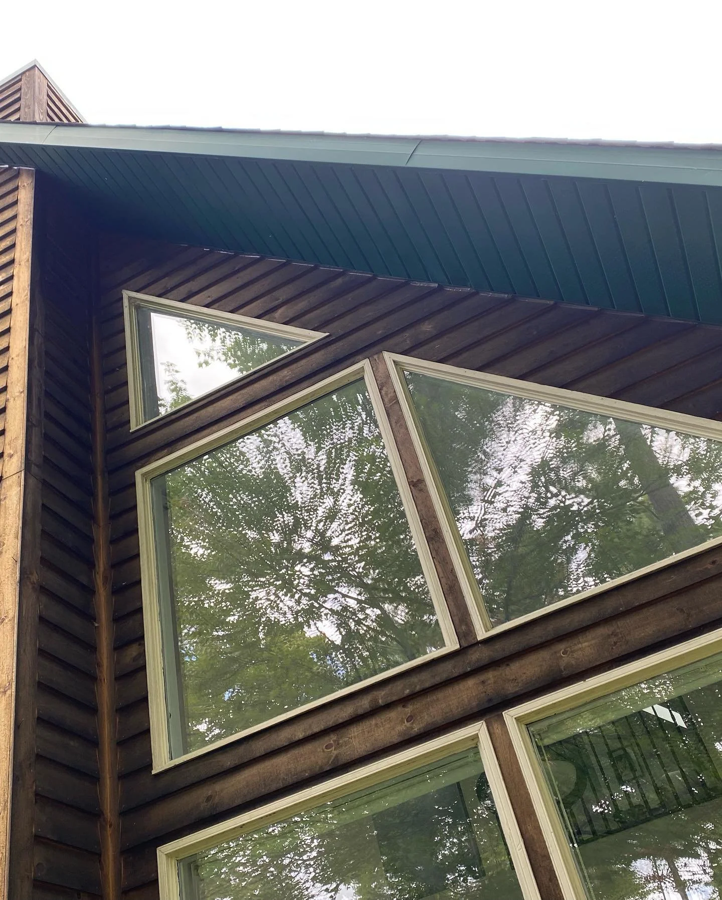 Close-up of a modern house with large angled glass windows reflecting trees, with dark wooden siding and a green roof.