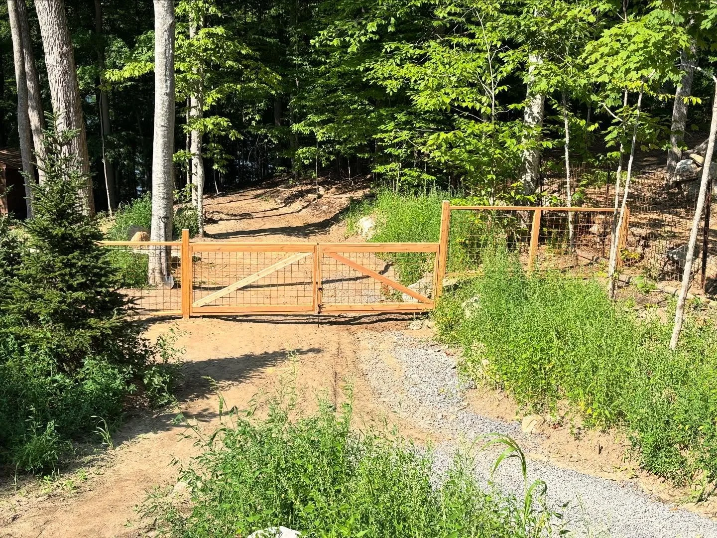 Wooden gate blocking a gravel and dirt path in a forested area with green trees and bushes on both sides.