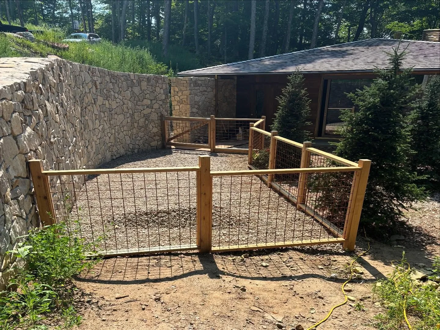 A fenced outdoor area with a gravel ground, surrounded by a stone retaining wall on one side and a wooden and wire fence on the other. There are small evergreen trees along the fence, and a building with a sloped roof in the background. A yellow hose