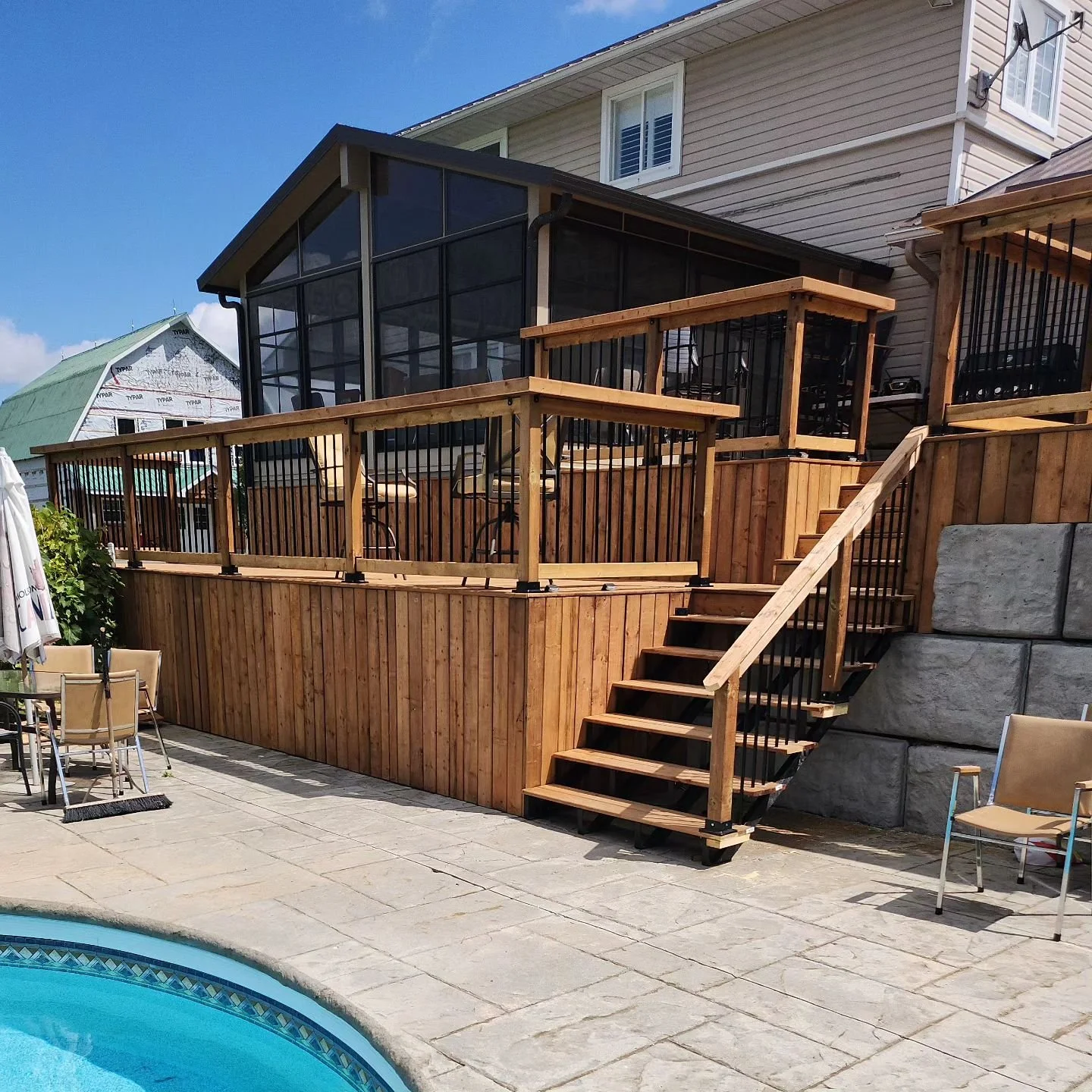 Wooden deck with stairs and railing, attached to a house, overlooking a pool in a backyard.