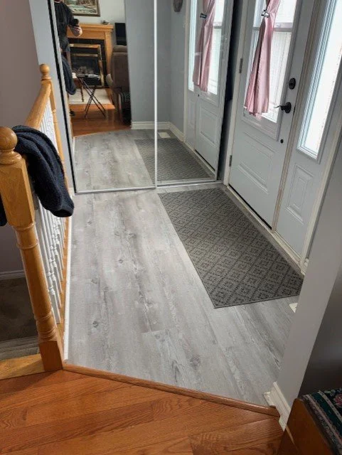 View of a home entryway with gray flooring, a mirror closet door, and a main door with pink curtains, a beige tiled floor, and a wooden staircase railing visible.