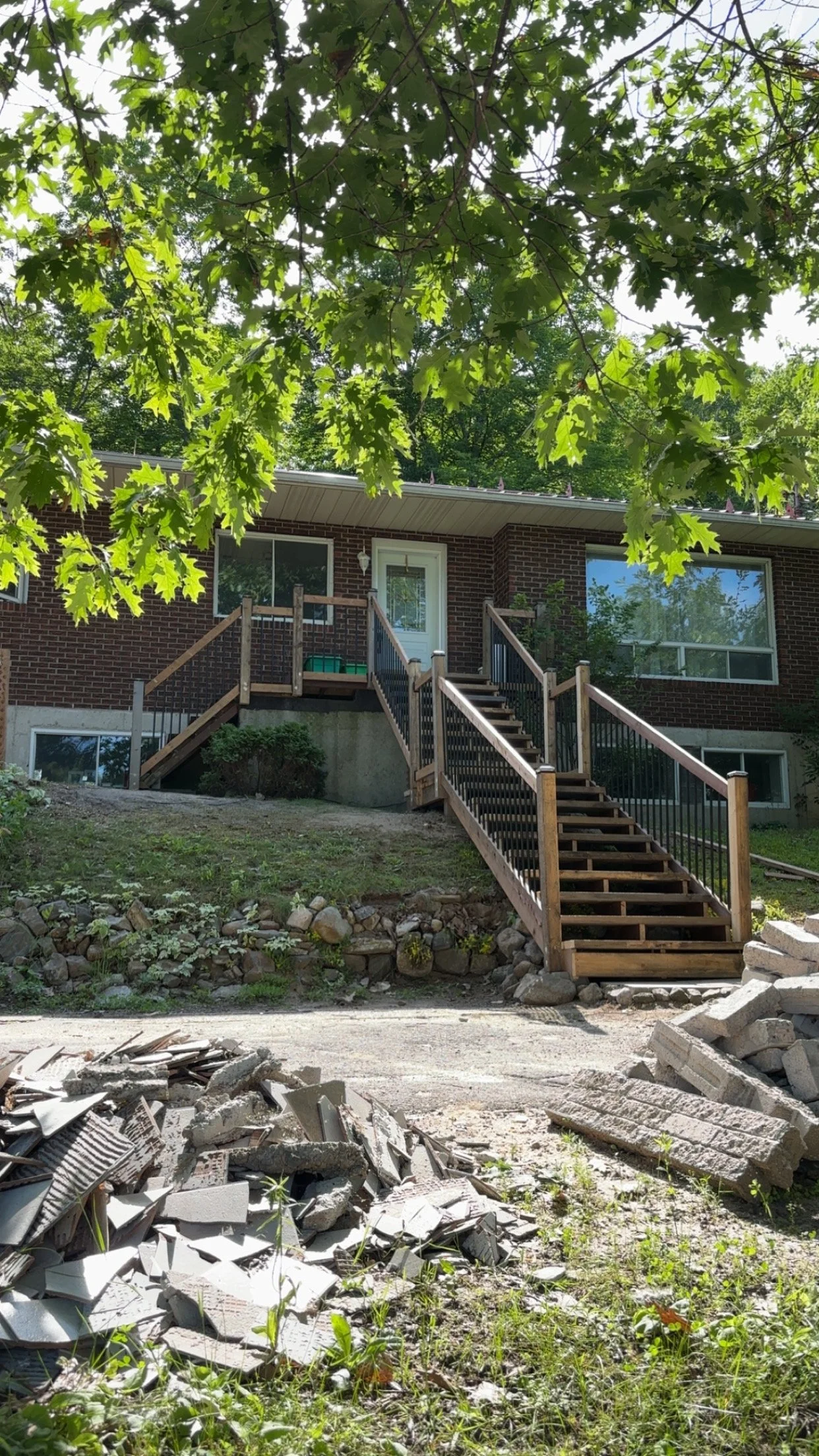 A brick house with a wooden staircase leading up to a front door, partially obscured by tree branches and leaves, with construction debris and rubble in the foreground.