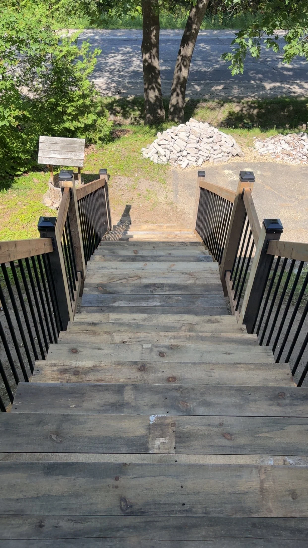 Wooden stairs with black metal railings leading down to a patch of grass and a sidewalk, with two piles of bricks and green trees in the background.