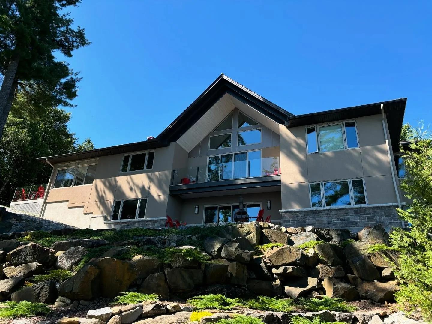 Modern house with large windows and a balcony, situated on a rocky hillside surrounded by trees, under a clear blue sky.