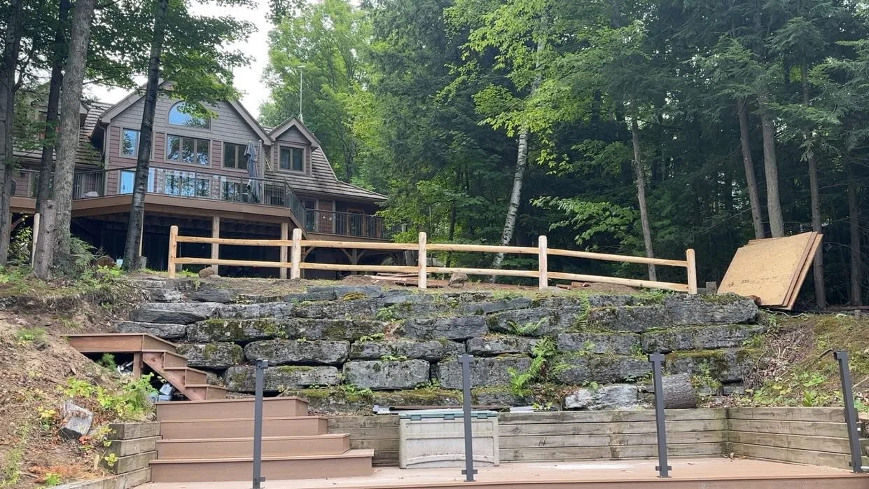 Construction of a new retaining wall and fencing around a sloped backyard with trees and a house in the background.