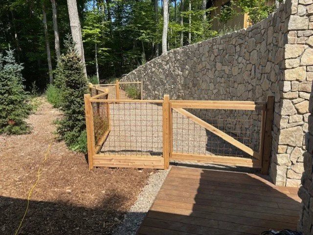 Wooden fenced gate with mesh panels leading from a wooden deck to a landscaped yard with trees and a stone retaining wall.