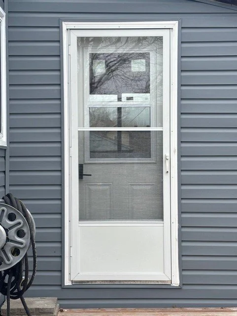 A white screen door installed on a blue house with grey vinyl siding. A garden hose is coiled on a reel to the left of the door.