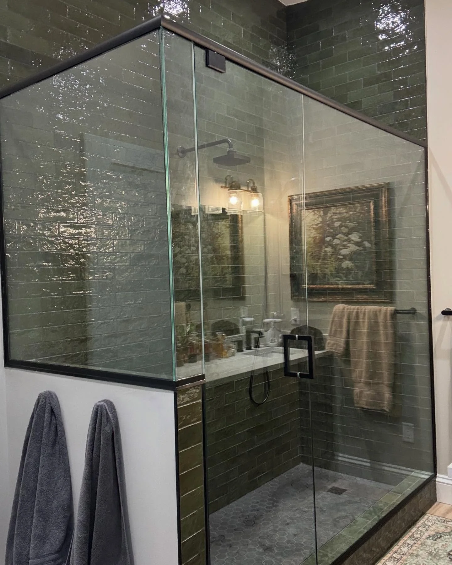 A modern bathroom shower with glass walls and dark green subway tiles, a black metal shower head, and a white vanity with toiletries reflected in the glass.