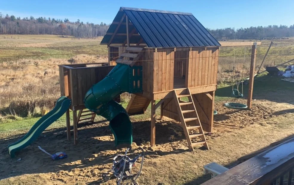 A wooden playhouse with a blue metal roof in a sandy yard. It has a green spiral slide and a straight slide, accessible by a wooden ladder. There are swings hanging from a nearby pole, and a child's stroller and other toys on the ground.