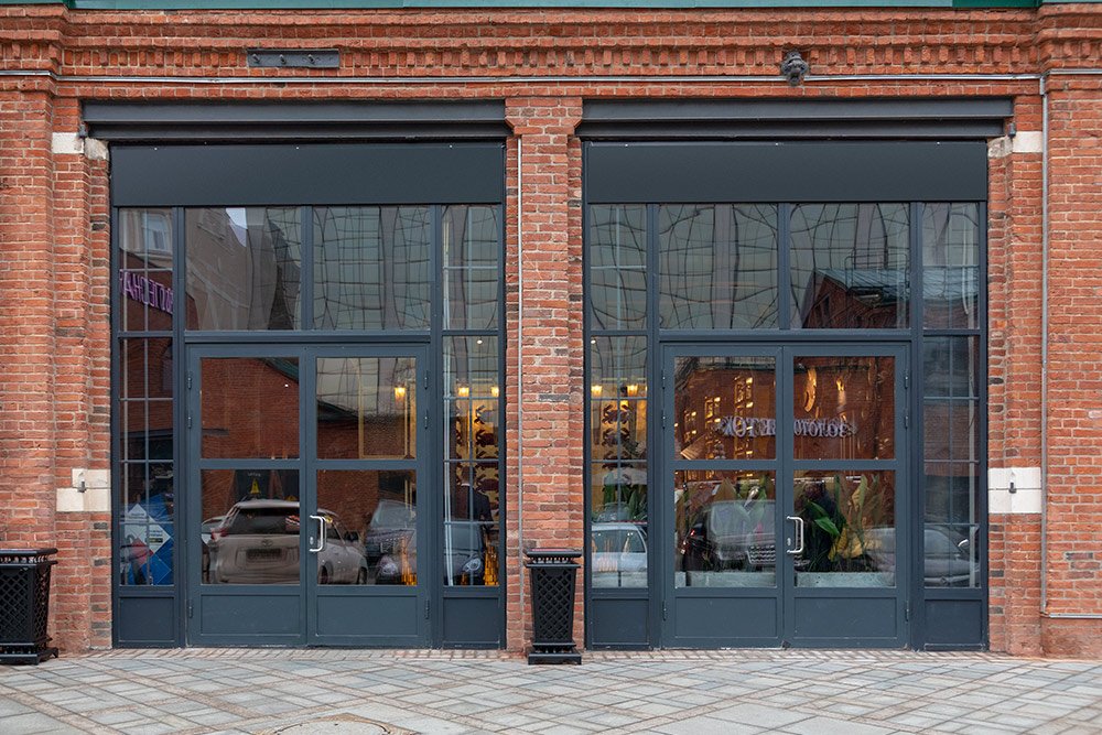 Two large glass storefront doors with dark frames on a brick building, reflecting the street outside and showing a glimpse of the interior.