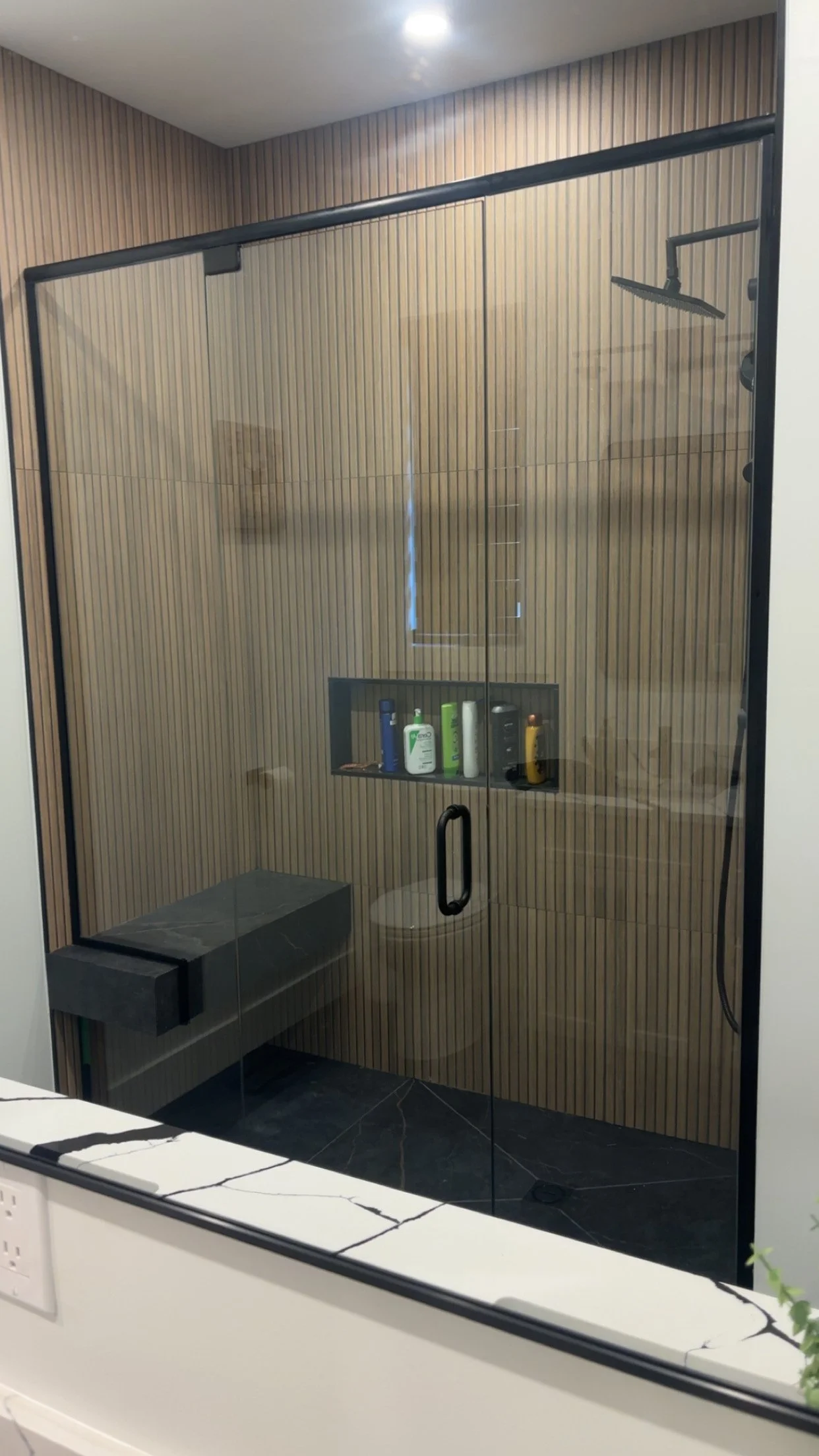 A modern shower with clear glass walls, wood-paneled walls, a rainfall showerhead, and various toiletries on a built-in shelf. There is a bench and a toilet inside the shower.