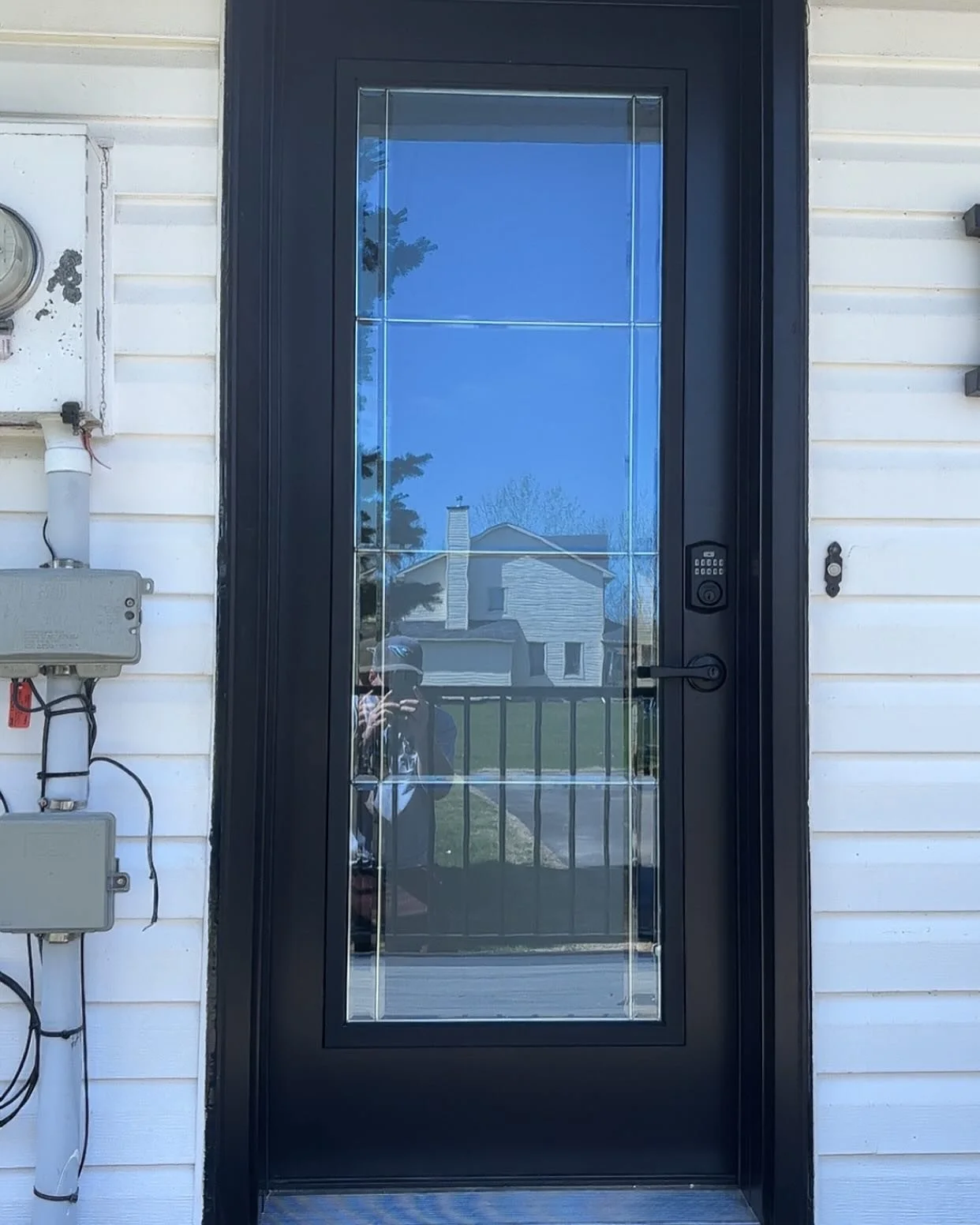Front door of a house with a black frame and a glass panel, reflecting the neighborhood and person taking the photo, mounted on white siding with visible utility boxes on the side.