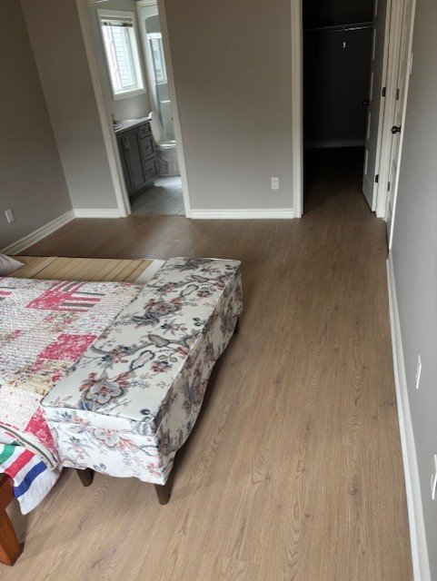 Empty bedroom with light gray walls, hardwood floor, bed partially visible with floral bedspread, an open closet, and a bathroom in the background.