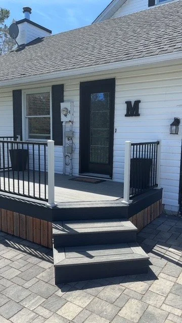 Front porch of a white house with black trim, featuring a small deck with black railings, three steps, a black door, a mailbox, a letterbox, and a large letter 'M' on the wall.