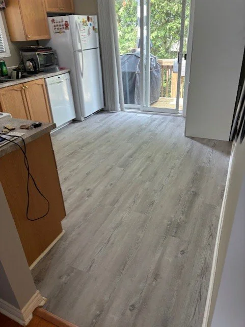 Empty kitchen with light wood flooring, white refrigerator with magnets, microwave, and a sliding glass door leading to a deck with a grill outside, and part of a wooden kitchen island.