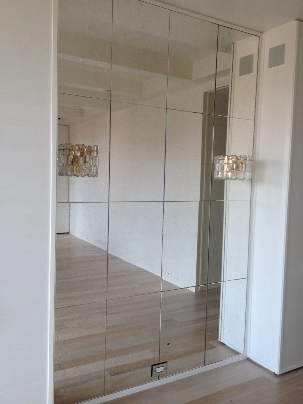 A large floor-to-ceiling mirror with a grid pattern in a modern room, reflecting a chandelier and part of a doorway.
