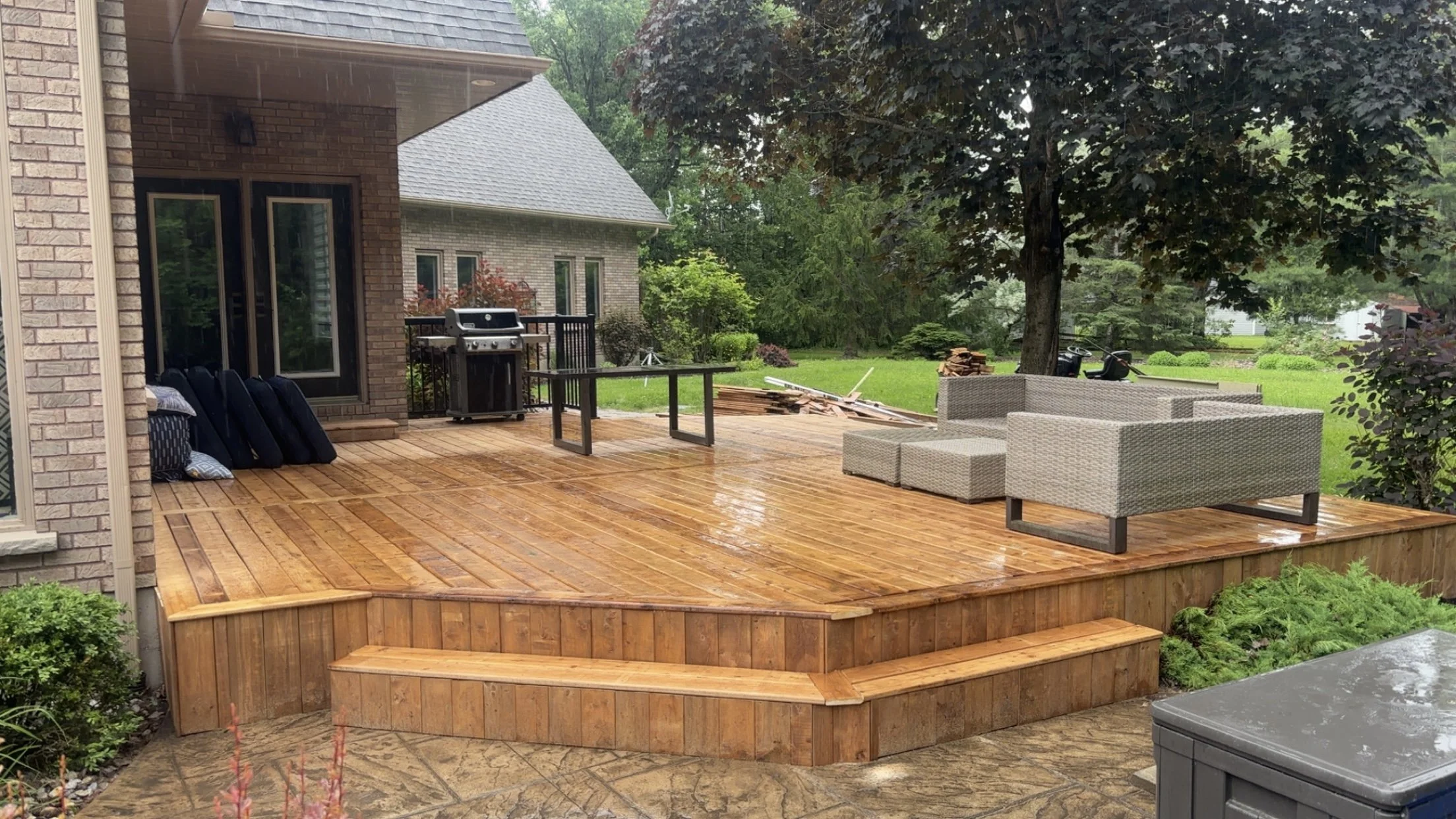 Rain-soaked wooden deck with outdoor furniture, a barbecue grill, and lush green trees and grass in the background.