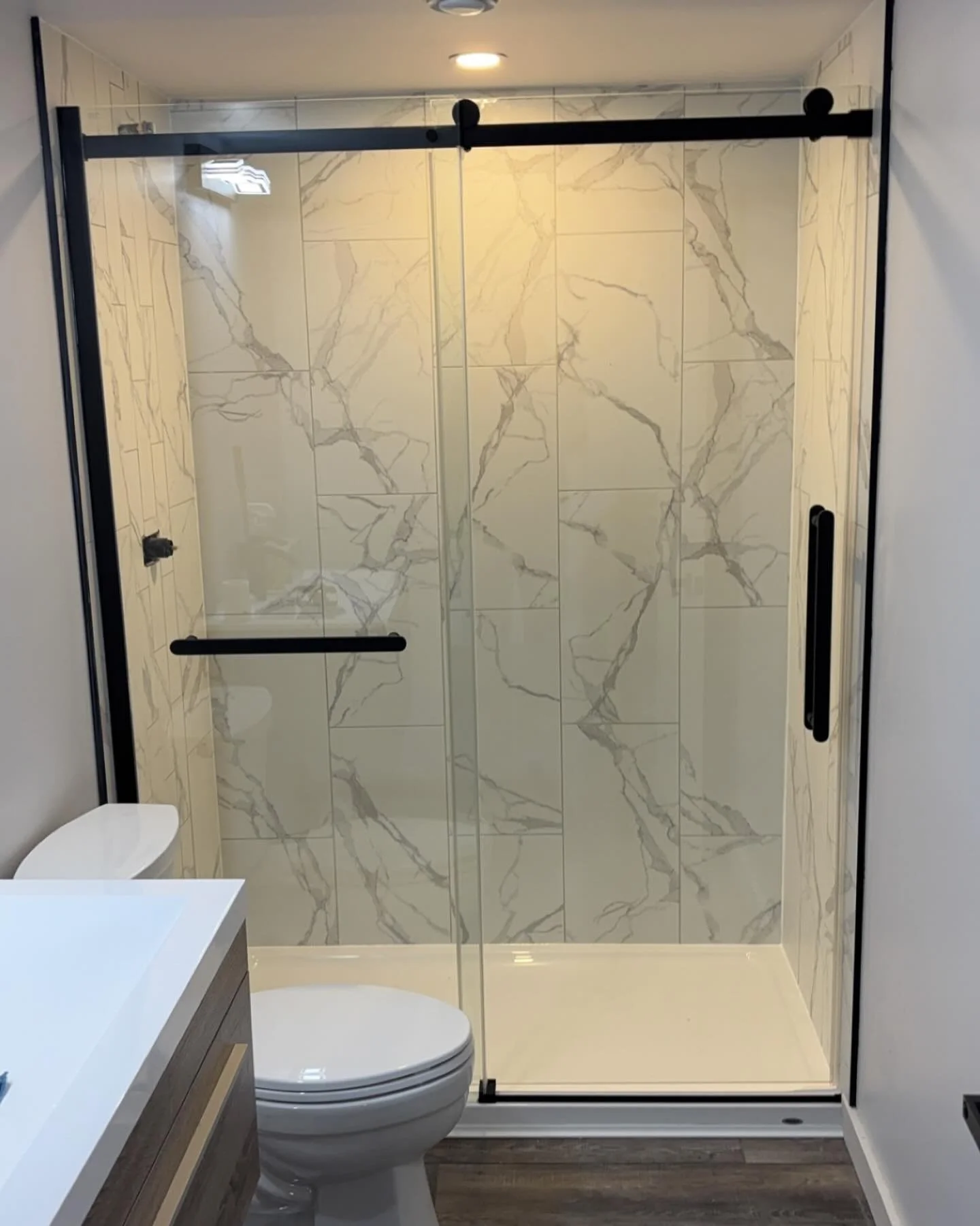 A modern bathroom featuring a walk-in shower with marble tile walls, a glass sliding door, a white toilet, and a wooden vanity with a white countertop.