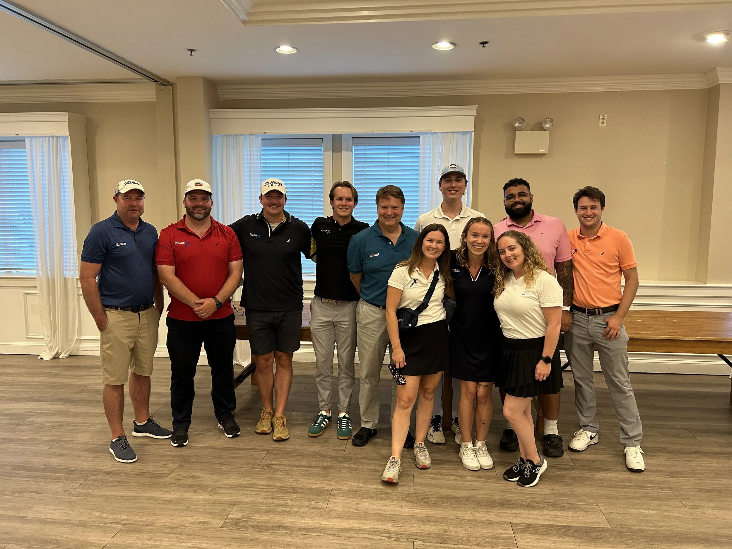 A group of twelve people, including men and women, standing inside a room with beige walls and wooden floors, posing for a photo. Some are wearing golf shirts, hats, and casual attire.