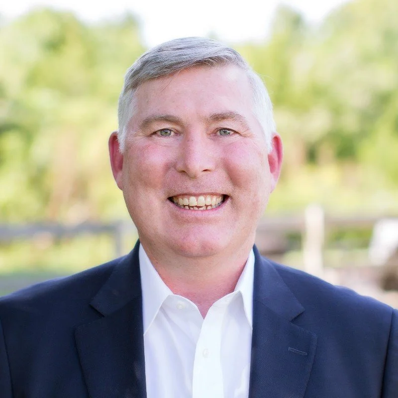 A smiling man dressed in a white shirt and navy blazer outdoors with green trees and blurred background.