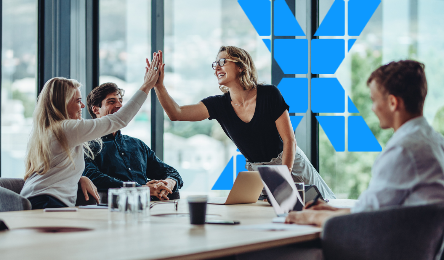 A group of four young professionals in a business meeting, with two women giving each other a high-five and smiling, in a modern office with large windows and a blue geometric wall design.