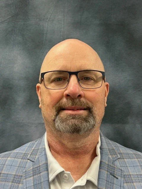 A bald man with glasses and a beard wearing a light blue plaid blazer and a white collared shirt, standing against a gray background.