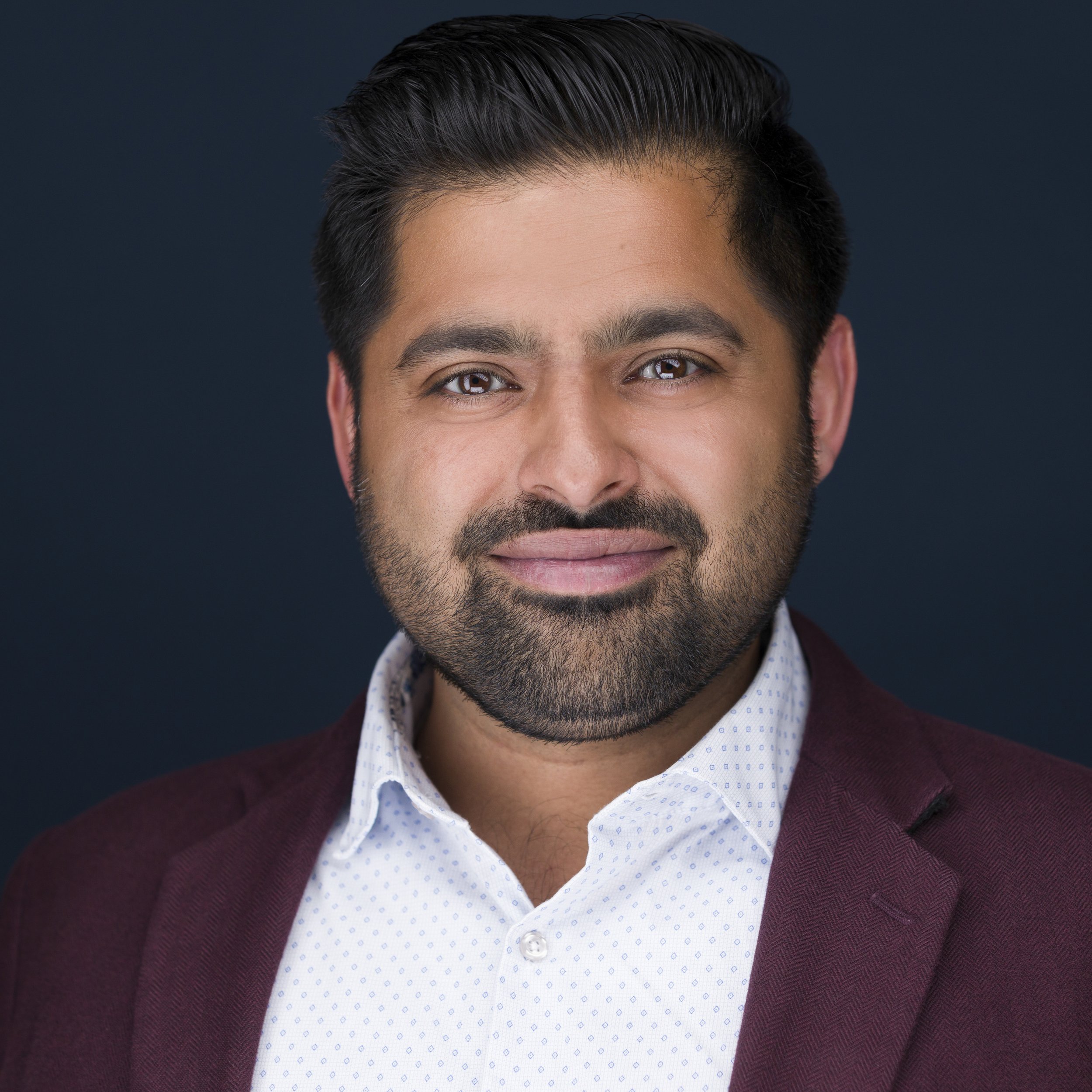 Headshot of a man with dark hair and beard, wearing a white shirt and a burgundy blazer, against a dark background.