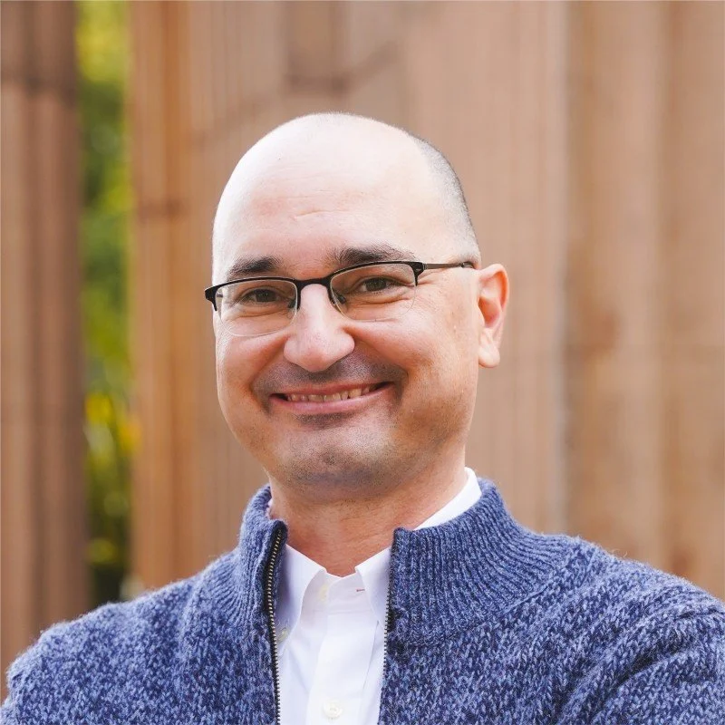 A smiling man with glasses, a shaved head, wearing a white collared shirt and a blue zip-up sweater, standing outdoors in front of a wooden fence.
