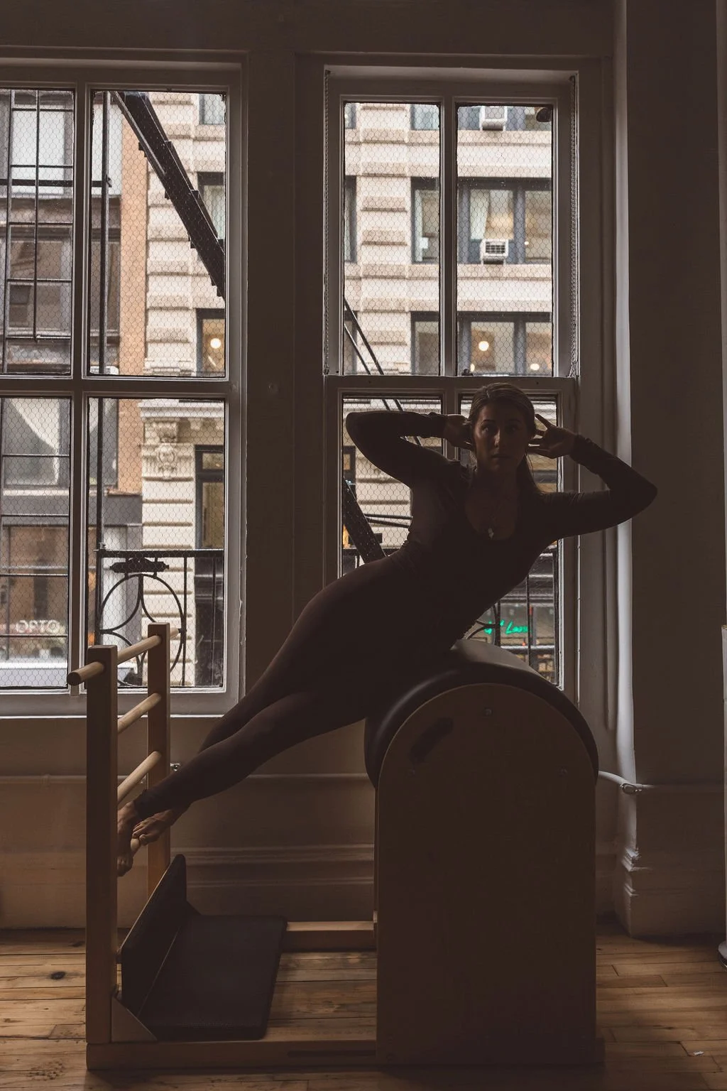 A woman in a dark outfit lying sideways on a piece of fitness equipment, with her head tilted back and hands behind her head, inside a room with large windows and an urban view outside.