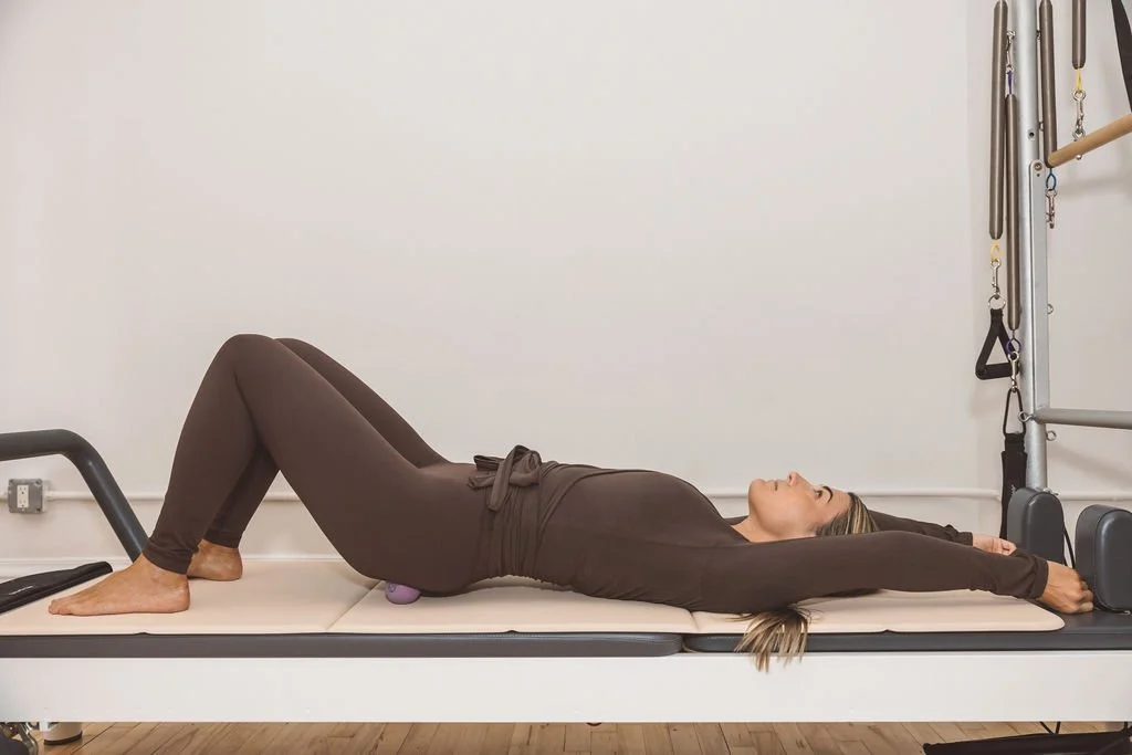 Woman lying on her back on a padded table during a Pilates or physical therapy session, with her arms stretched above her head, holding onto Pilates equipment.