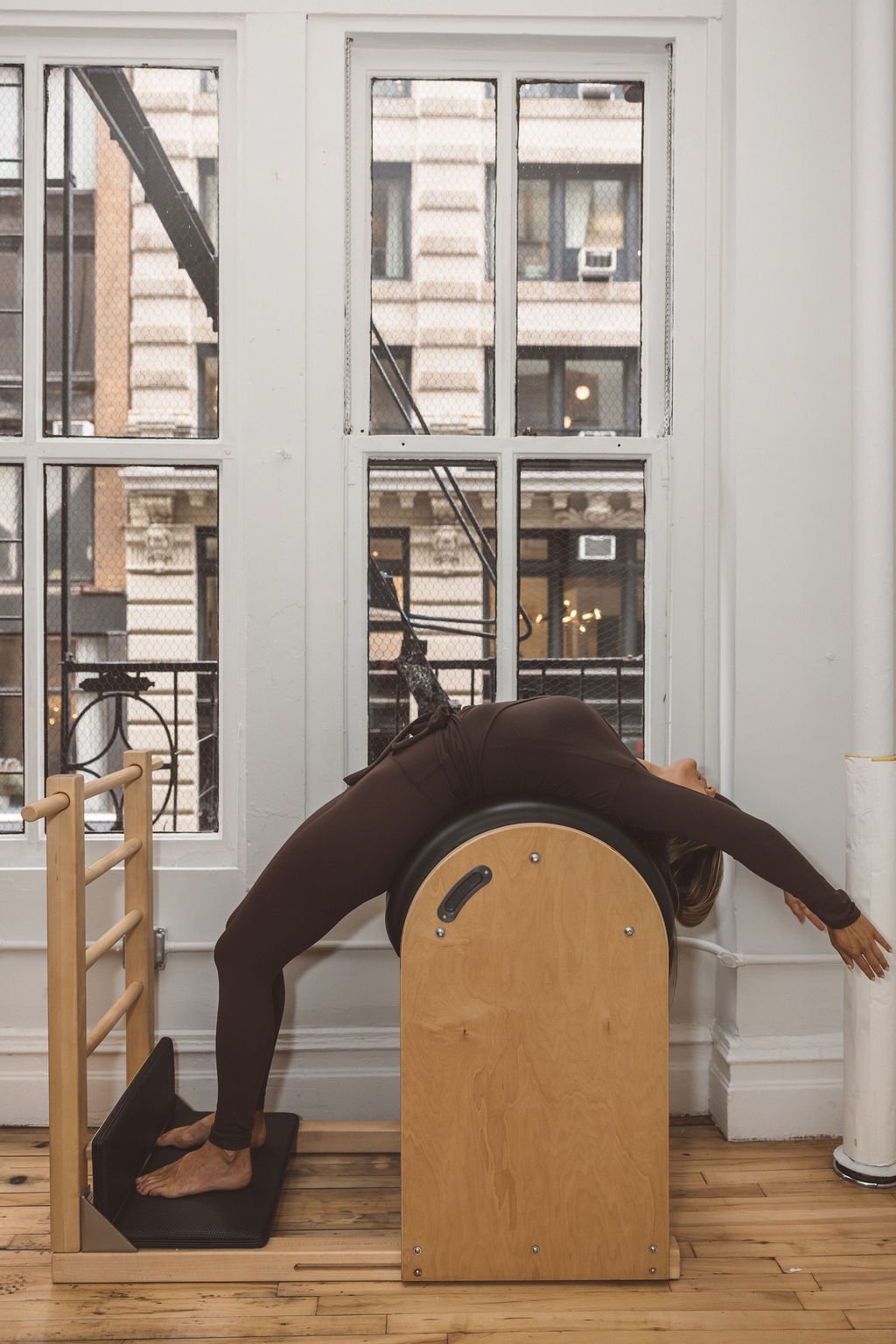 A woman is performing a backbend stretch on Pilates equipment in a studio, with large windows and city buildings in the background.