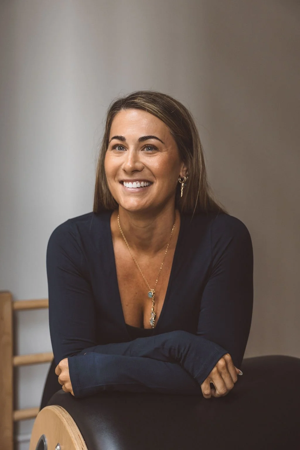 A woman with long brown hair smiling, wearing a black top, gold jewelry including earrings and a necklace, sitting with her arms crossed on a black object, against a plain beige wall background.