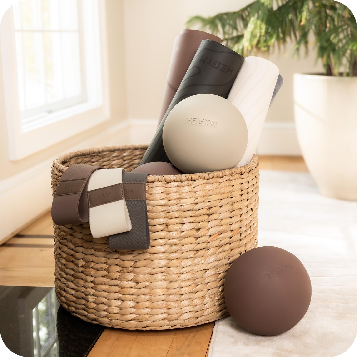 A woven basket filled with fitness accessories including mats, a ball, and resistance bands, placed on a wooden table in a bright room with a large window and a potted plant in the background.