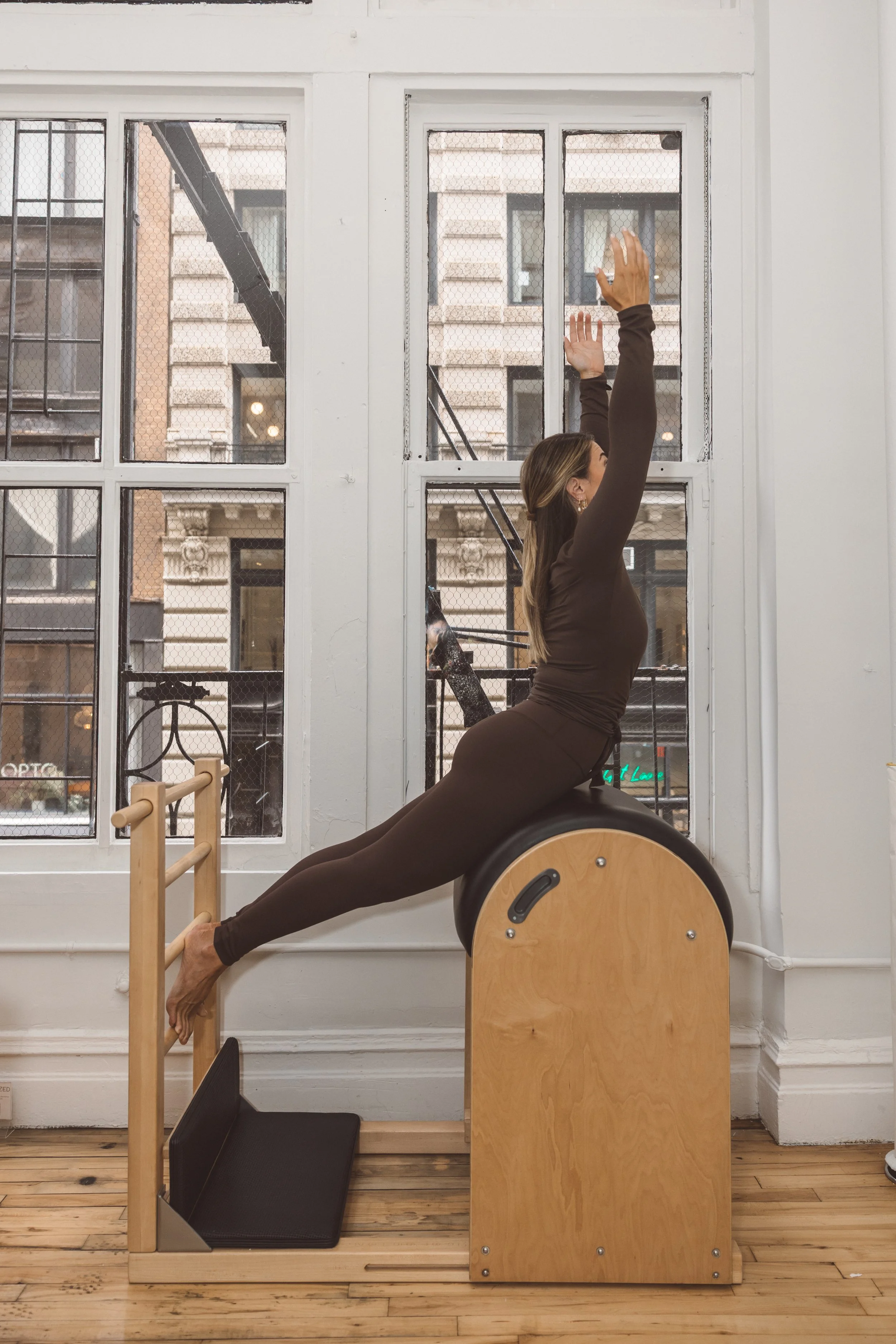 A woman practicing Pilates on a specialized chair with her back arched and arms extended upward, in a bright studio with large windows showing an urban cityscape outside.